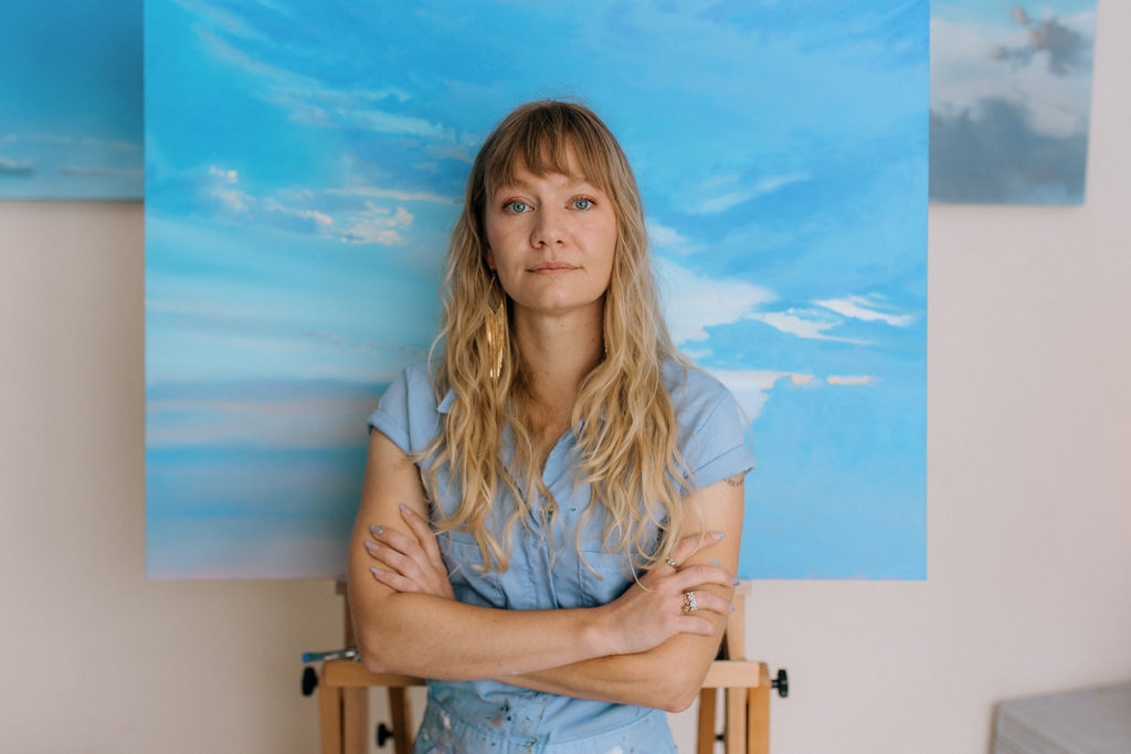 Anneio Klaas photographed in front of oil painting of skyscape.