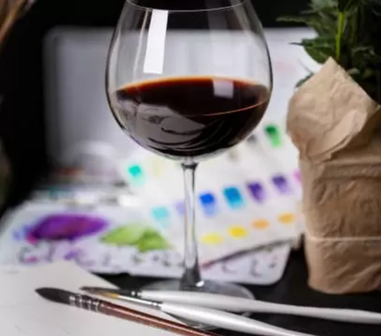 Glass of red wine, paint pallets of greens, blues, and purples, and paintbrushes